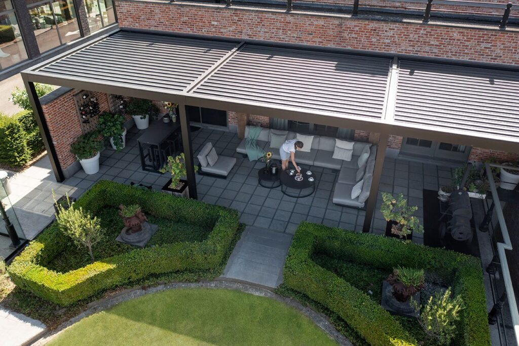 Ruim en modern terras onder een Harol terrasoverkapping met lamellen, voorzien van een grijze loungeset en eettafel, omringd door strak gesnoeide hagen. Een persoon dekt de tafel, terwijl de overkapping zorgt voor schaduw en comfort tegen een bakstenen gevel.